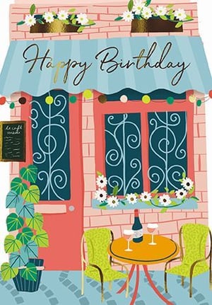 a happy birthday card with a brick building with a table and chairs in front of it .