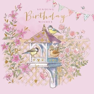Send warm birthday wishes with this enchanting card, perfect for any special woman in your life. Featuring a delightful garden scene, it showcases a charming bird feeder bustling with sweet blue tits and a tiny wren. Delicate pink clematis and climbing roses intertwine gracefully around a rustic trellis, creating a serene backdrop. Elegant bunting flutters above, adding a touch of celebratory charm. With "Sending Birthday Wishes" beautifully scripted in shimmering gold, this card offers a gentle and heartfelt way to celebrate her day. Its soft pastel hues and intricate artwork make it a truly lovely choice.